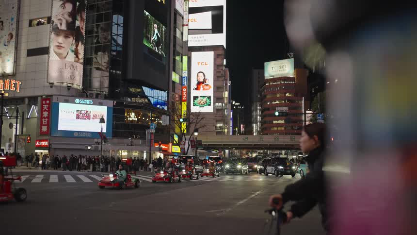 A slow-motion view of go-kart tourists driving through the iconic Shibuya Crossing at night, surrounded by bright city lights and traffic