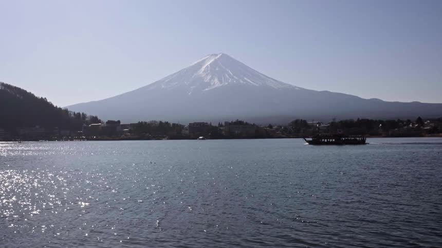 A tour boat slowly crosses a shimmering lake with the iconic, snow-capped Mount Fuji in the background on a sunny day