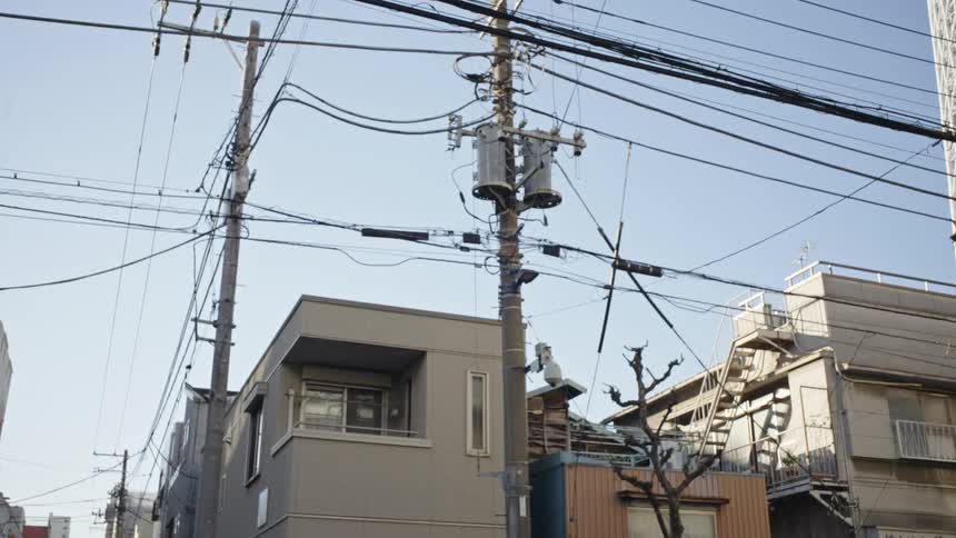 A complex web of power lines and utility poles crisscrosses the clear sky above a quiet Japanese residential street