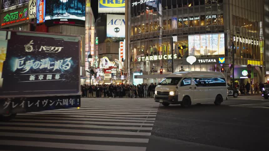 An anime advertisement truck drives through the iconic Shibuya Crossing at night in slow motion, surrounded by neon lights