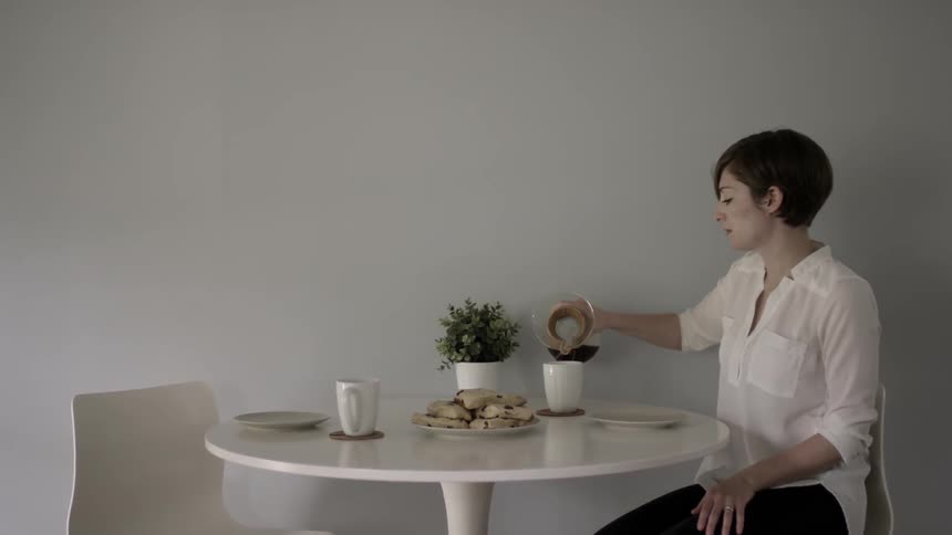 A woman sitting down a pastry on a table and then commencing to pour coffee into her mug