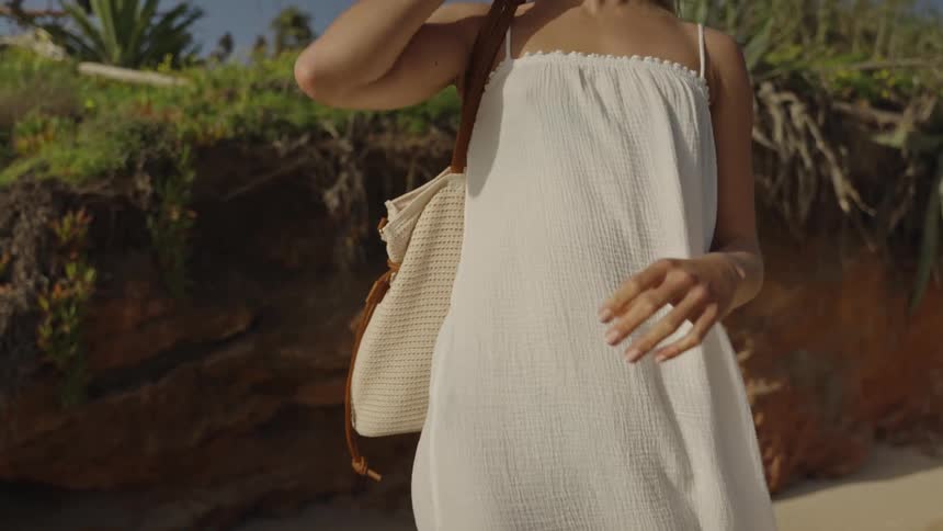 A woman in a white sundress adjusts her woven beach bag on her shoulder on a sunny day