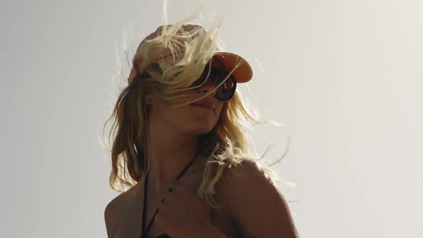 A stylish blonde woman in a cap and sunglasses enjoys the summer breeze as her hair blows in slow motion