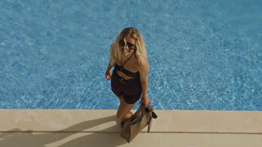 A high-angle shot of a stylish woman with dwarfism smiling up at the camera before walking along the edge of a sunny swimming pool