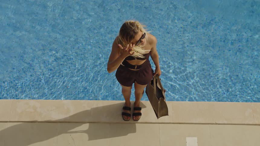 Slow motion high-angle shot of a fashionable woman standing at the edge of a bright blue swimming pool on a windy day