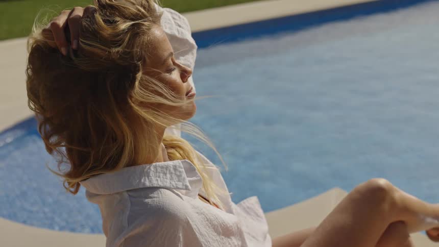 A blonde woman relaxes by the pool in slow motion, running her hand through her hair as it blows in the summer breeze