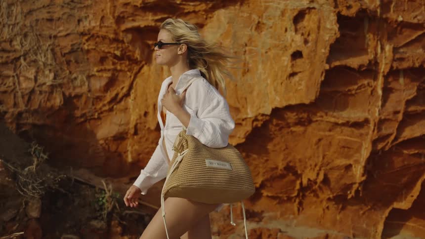 A stylish blonde woman walks in slow motion along a beach with a straw bag and sunglasses, her hair blowing in the wind in front of a stunning red rock cliff
