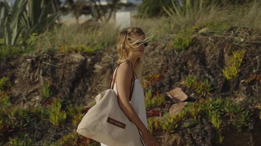 Slow motion shot of a fashionable woman in sunglasses and a white dress walking along a verdant sand dune path