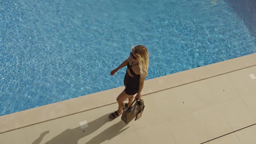 An elegant blonde woman in sunglasses walks in slow motion along the edge of a beautiful swimming pool at a luxury villa