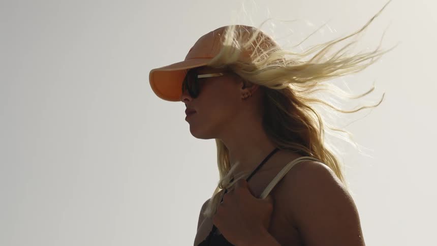 A low-angle, slow-motion shot captures a stylish blonde woman in a cap and sunglasses as her hair whips in the wind