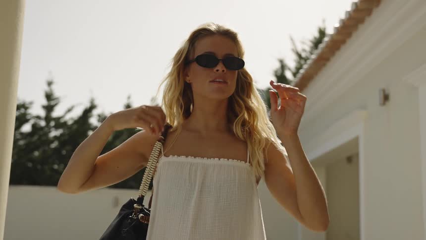 A stylish blonde woman in a white dress slowly puts on her sunglasses while enjoying a sunny day at a resort villa