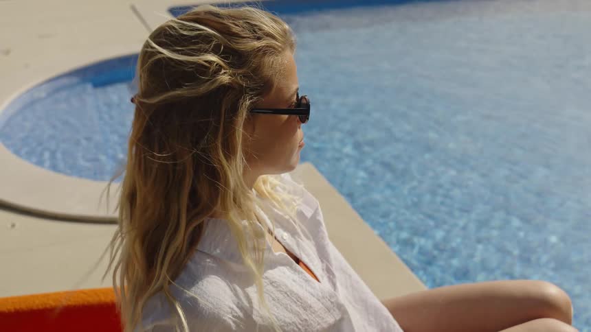 A beautiful blonde woman relaxes in the sun by the pool as the wind blows through her hair in slow motion