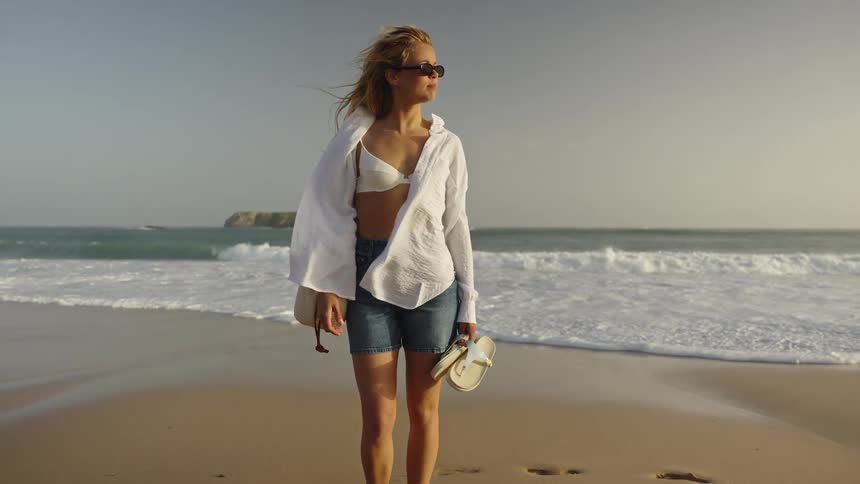 A cinemagraph of a stylish blonde woman standing on a windy beach and looking out at the ocean waves during golden hour