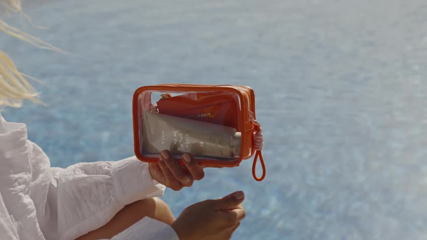 A woman in a white shirt holds and unzips her clear, orange cosmetic bag full of beauty products while sitting by a sunlit pool