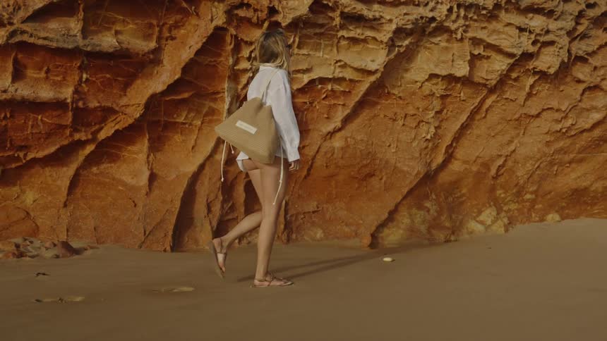 A woman in a white shirt and sandals walks in slow motion along a sandy beach next to a large orange rock formation