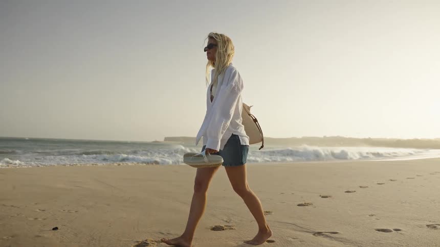 A stylish blonde woman enjoys a peaceful, barefoot walk along a sandy beach at golden hour, carrying her sandals