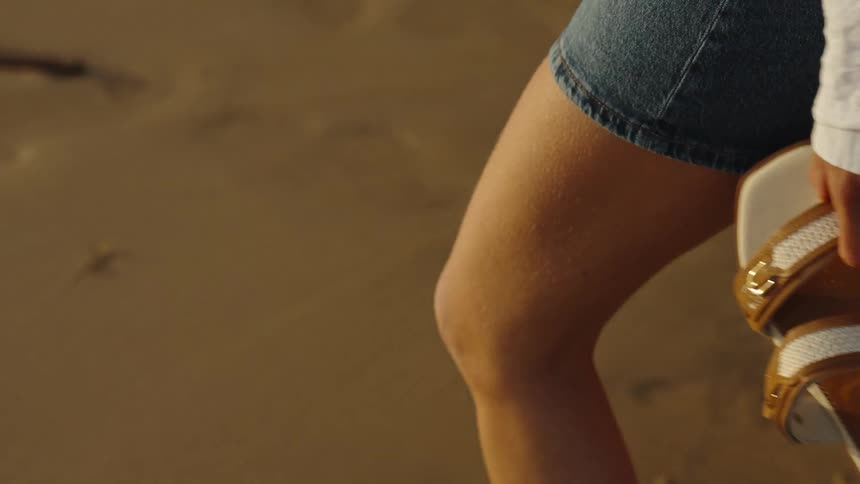 A woman in denim shorts and a white shirt walks along a sandy beach in slow motion, carrying her sandals