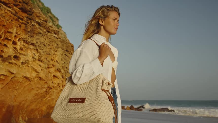 A stylish blonde woman with a woven shoulder bag stands on a beach near golden cliffs, enjoying the serene ocean view at sunset