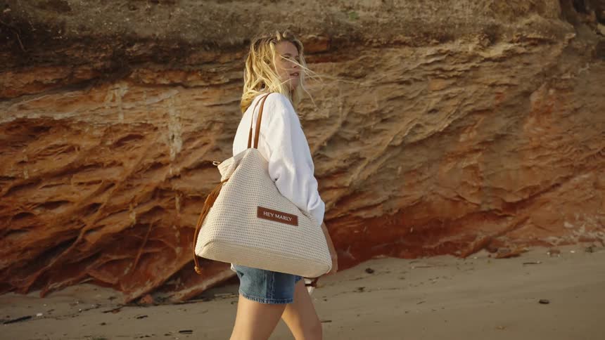 A stylish blonde woman walks in slow motion along a sandy beach beside a red rock cliff, carrying a tote bag
