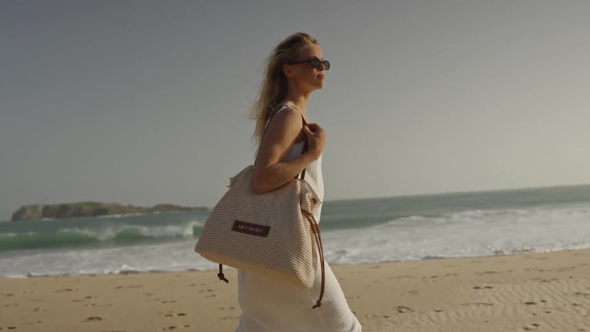 A blonde woman in a flowing white dress and sunglasses enjoys a peaceful, windy walk along the sandy ocean shoreline
