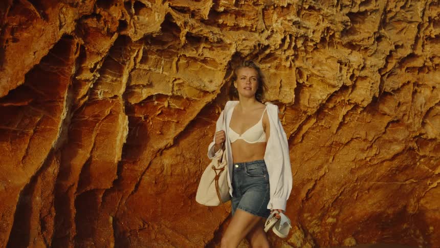 A windswept blonde woman enjoys a serene walk barefoot on the wet sand beside dramatic golden rock formations at sunset
