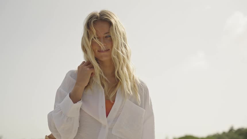 A beautiful blonde woman with windblown hair puts on her beach bag and smiles warmly at the camera in slow motion