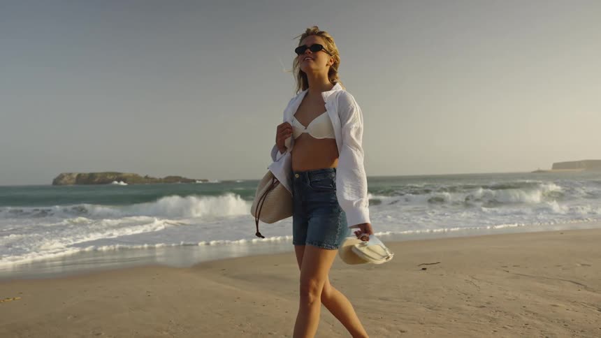 A fashionable blonde woman in a bikini and jean shorts walks barefoot in slow motion along a sandy beach with a tote bag and sandals in hand