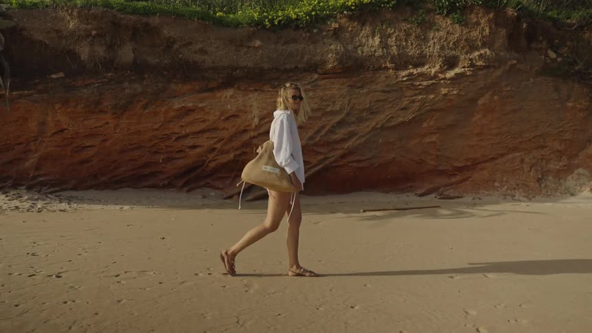 A stylish woman in a white shirt walks in slow motion along a sandy beach next to a cliff, carrying a large woven bag