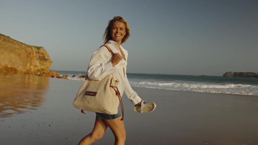 A smiling blonde woman walks barefoot along the shoreline at sunset, carrying her bag and sandals as the waves gently wash ashore