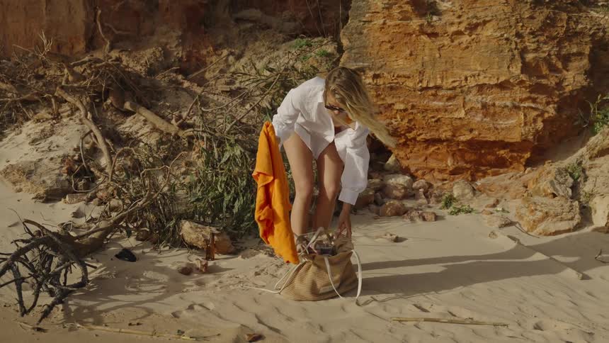 A blonde woman walks along a sandy beach, then stops to lay out her bright orange towel by a rocky cliff to sunbathe