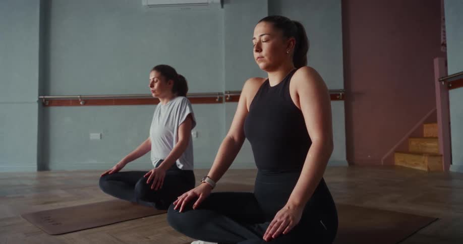Two serene women with eyes closed practice deep breathing meditation while sitting on mats in a tranquil yoga studio