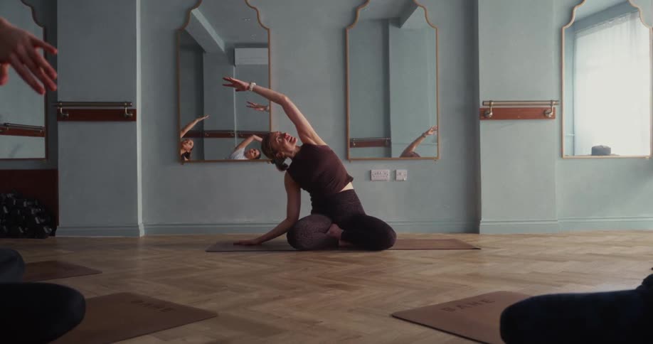 A woman gracefully performs a seated side bend yoga stretch on her mat during a class in a serene studio