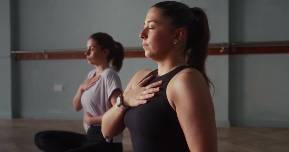 Two women practice calm, meditative breathing with their eyes closed in a quiet fitness studio