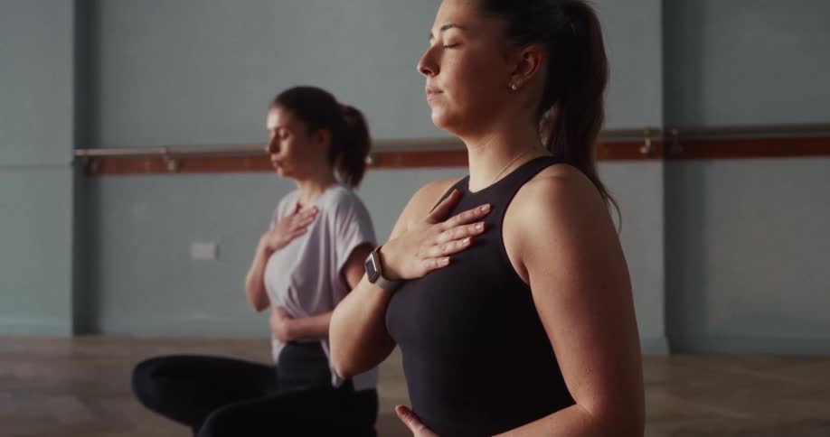 Two serene women with eyes closed practice deep breathing and meditation while sitting on mats in a tranquil yoga studio