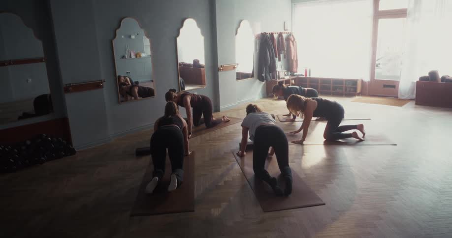 In a serene, sun-drenched studio, a group of women gracefully transitions from child's pose to tabletop position during a yoga session