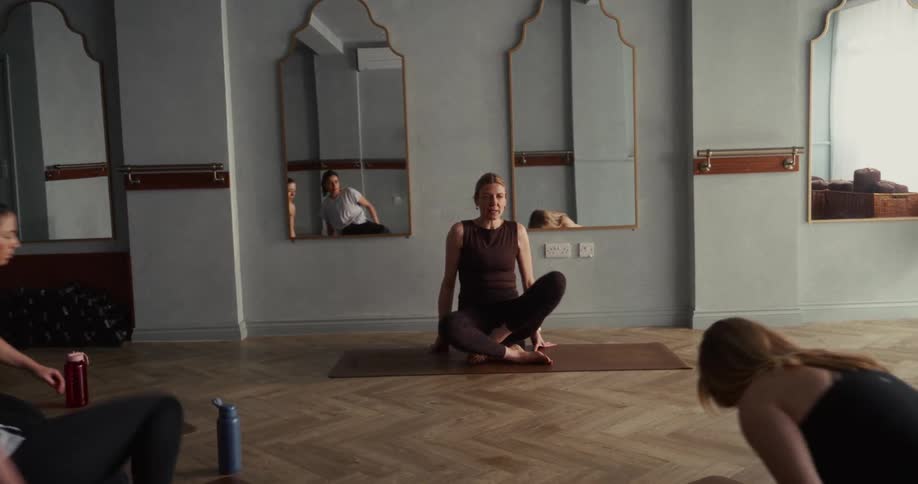 A female yoga instructor demonstrates a flow and sits in a meditative pose for her class in a tranquil studio