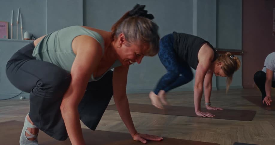 Two women in a yoga class transition through a challenging arm balance sequence in slow motion