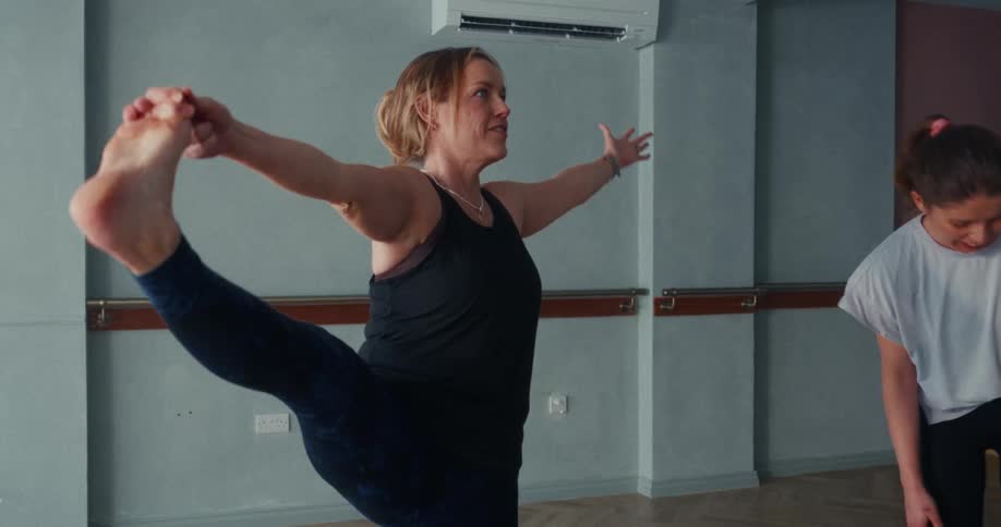 A woman in a yoga class attempts a balancing pose, wobbles, and then laughs as she falls out of the position
