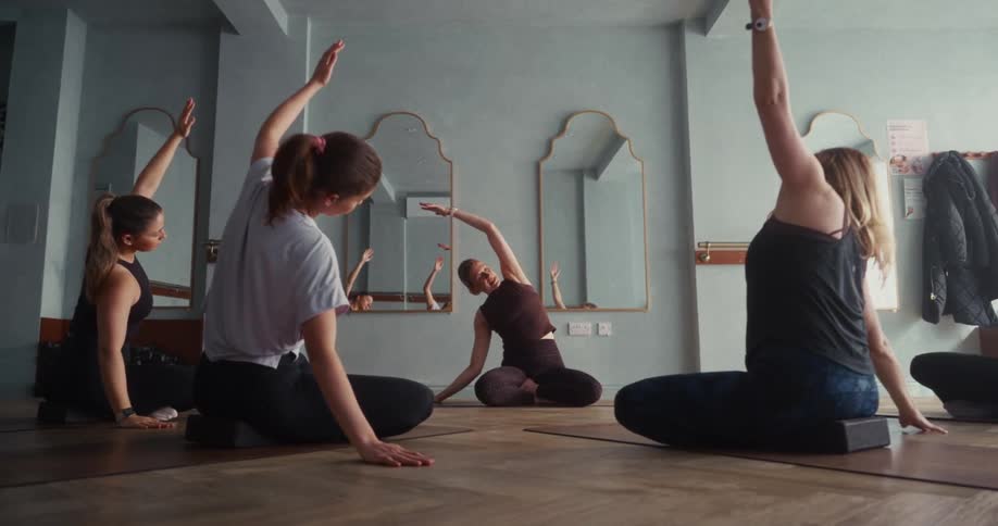 A group of women follow their instructor's lead in a seated side stretch during a calm yoga class
