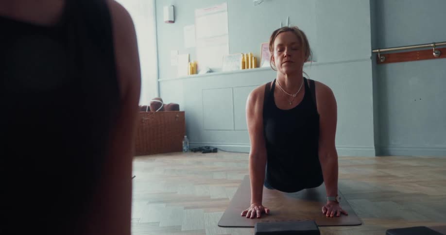 Slow motion reveal of a focused woman transitioning into an upward-facing dog yoga pose in a fitness class