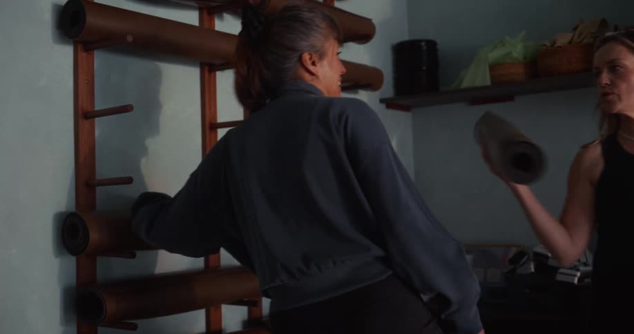 A woman in workout clothes takes a rolled-up yoga mat from a wall-mounted rack in a studio