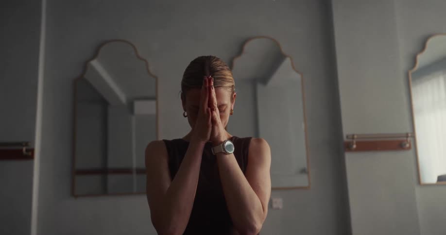 A calm woman meditates with her hands in a prayer pose inside a serene, dimly lit studio