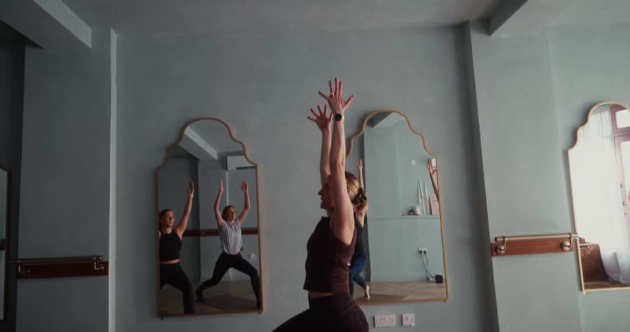 A woman holds a high lunge yoga pose with her arms raised during a class in a serene studio, captured in a slow upward tilting shot