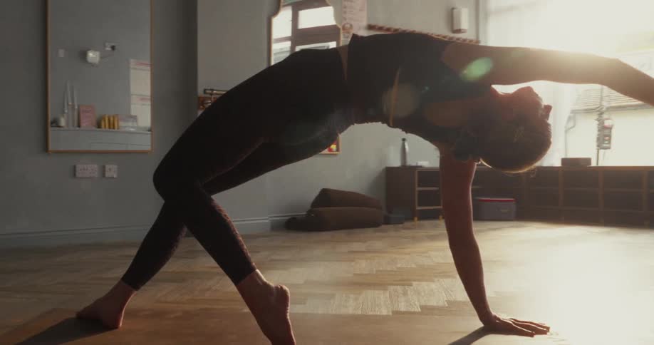 A graceful woman holds a challenging backbend yoga pose on her mat in a sunlit studio in slow motion