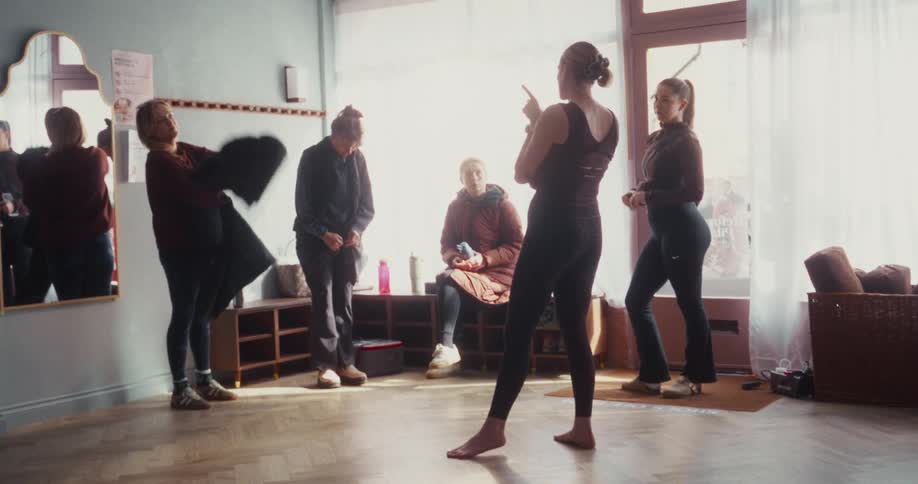 A group of women arrives and prepares for a yoga class, chatting with the instructor in a sunlit studio