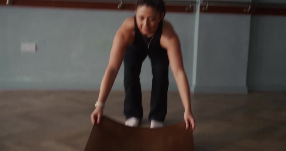 A fit woman in slow motion unrolls a brown yoga mat onto the wooden floor of a fitness studio
