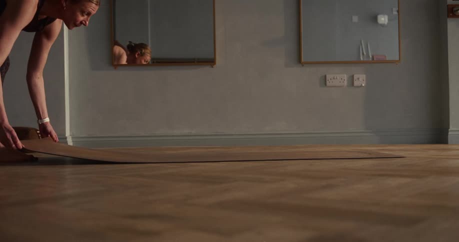 In slow motion, a woman unrolls a yoga mat across a wooden herringbone floor in a tranquil studio