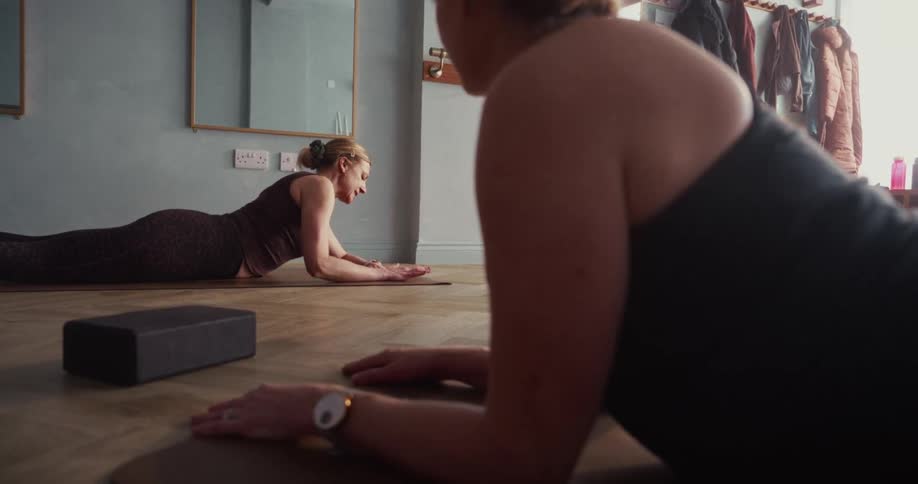 Two women in a bright studio gracefully transition from a yoga pose to lying on their mats in slow motion