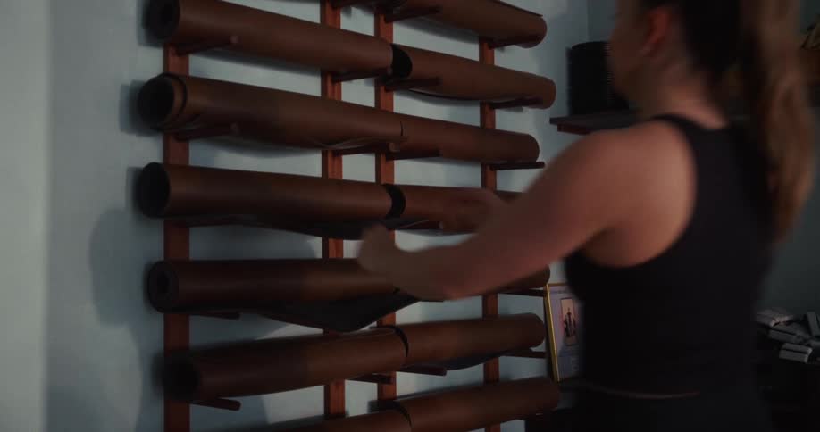 A young woman takes a brown yoga mat from a wall-mounted rack in a fitness studio