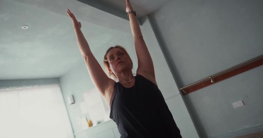 A determined blonde woman holds a warrior yoga pose with arms outstretched in a bright, modern studio